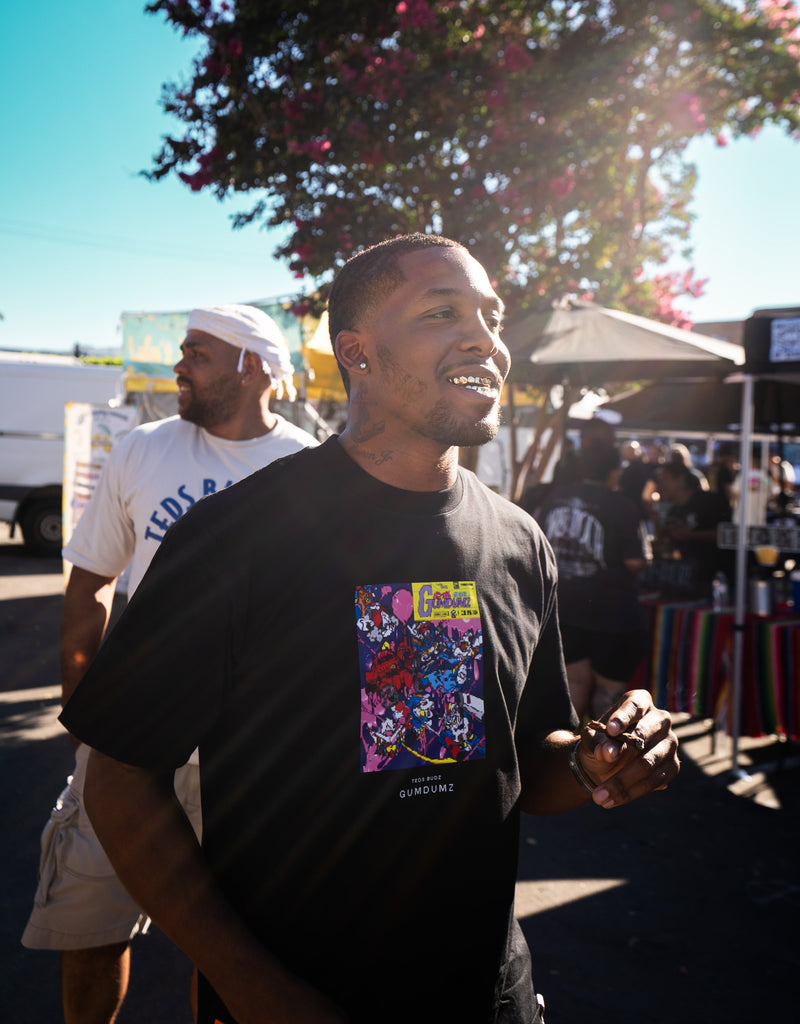 Man wearing a black t-shirt with colorful graphic design, standing outdoors with trees and people in the background.